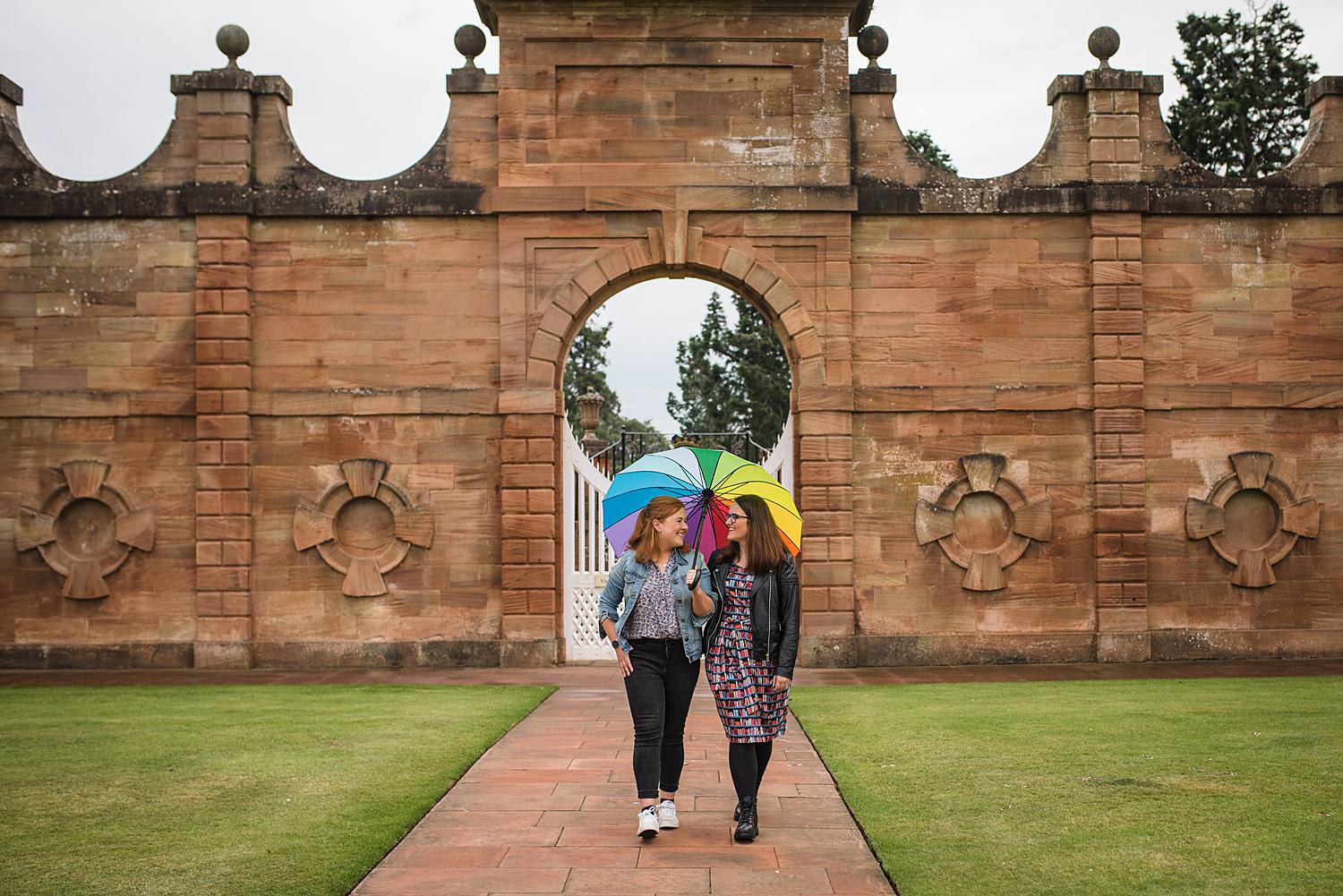 Two woman in love walk down a path at Chatelherault Hunting Lodge carrying a rainbow umbrella